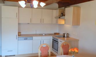 A modern kitchen with white cabinets and a wooden ceiling. A round dining table with chairs and a small flower vase is in the room.