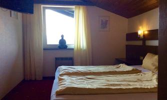 A cozy room with a double bed and a television. The room has large windows with curtains and a statue of a Buddha on the windowsill.
