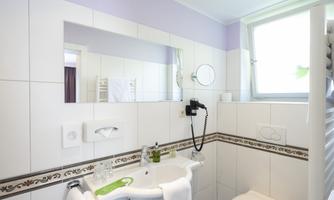 A modern bathroom with white tiles and a large mirror wall. There is a shower, a sink, and a window that lets in natural light.