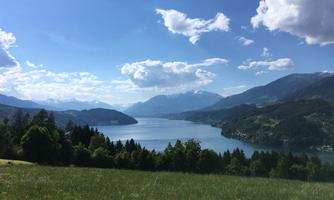 A picturesque lake surrounded by mountains and green forests. The sky is blue with white clouds.