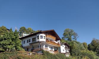 A picturesque house on a hill, surrounded by trees and plenty of green foliage. The sky is clear and sunny.