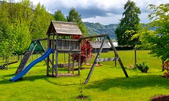 A playground with a wooden play structure and a blue slide. Surrounded by green grass and trees, it is ideal for children.