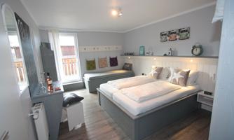 A cozy bedroom with two beds and a small sofa. There are large windows with natural light and a subtle, modern decor.