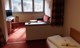 A cozy hotel room with a bed, a small table, and a television. Light streams through the windows, and there is an area with a couch and an armchair.