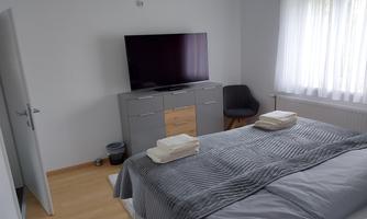 A modern bedroom with a large television and a comfortable bed. The room has large windows that let in plenty of daylight.