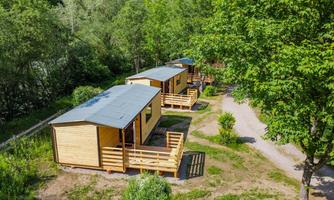 Three modern wooden holiday homes are located in a green environment. They have visible terraces and are surrounded by trees.