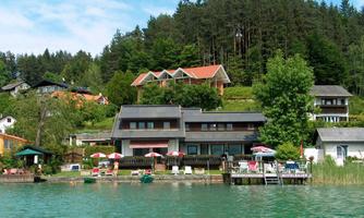 A picturesque shore with several houses and a beautiful view of the forest in the background. By the water, there are sun loungers and umbrellas arranged for relaxed days.