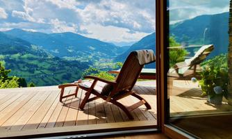 A cozy lounge chair on a terrace with a breathtaking view of green mountains and valleys. The clouds in the sky give the scene a relaxed atmosphere.