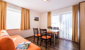 A bright room with an orange sofa and a dining area. Large windows with curtains provide plenty of daylight.
