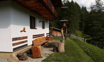 A rustic house with a wood stove and a seating area made of solid wood. In the background, trees and a meadow can be seen.