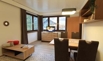 A modern living room with large windows and bright wooden furniture. The room has a dining table and a cozy seating area.