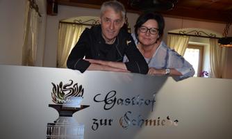 A friendly couple stands behind a sign that reads "Inn at the Forge." The background features a cozy interior.