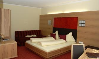 A modern hotel room with a double bed and a couch. The walls are designed in warm wood and red tones, with cozy lighting.