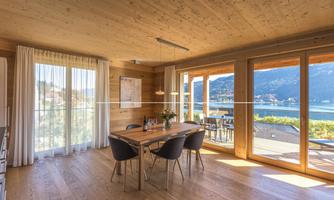 A bright dining room with wooden floors and large windows. The view looks out onto a lovely landscape with a lake and mountains.