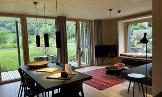 A modern living room with large windows overlooking the green landscape. The space is stylishly furnished with a dining table, sofa, and decorative elements.
