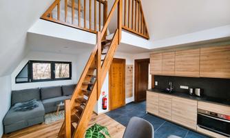 A modern living room with an open kitchen and a wooden staircase. The space is bright and welcoming with a cozy sofa and a beautiful plant.