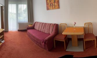 A cozy living room with a red sofa bed, a table and chairs. There is a television and a small kitchen in a bright room.