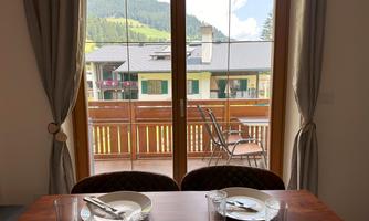 A dining area with a table set with dishes and glasses. Through large windows, there is a view of a green landscape with balconies.