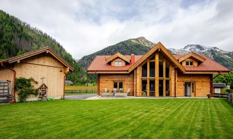 A modern wooden house with large windows and a red roof, surrounded by a green meadow. In the background, majestic mountains rise under a cloudy sky.
