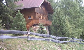 A rustic wooden house surrounded by green trees. In the foreground, there is a fence that encloses the property.