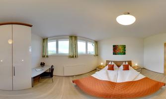 A bright bedroom with a large bed and orange bedding. The room has large windows and a desk in the corner.
