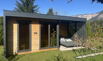A modern wooden house with large windows and a cozy outdoor area. Surrounded by a well-maintained garden and mountains in the background.