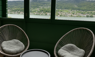 A cozy room with two stylish armchairs and a small table. Through the windows, a green landscape by the shore and a cloudy sky can be seen.