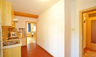 A bright kitchen with wooden shelves and a continuous hallway. The floor is tiled with terracotta-colored tiles.