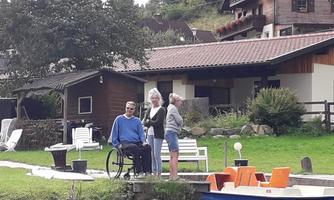 A man in a wheelchair is sitting by the shore of a lake and talking to two women. In the background, cozy wooden houses and a green landscape can be seen.