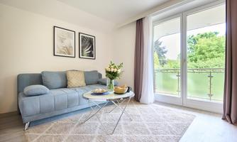 A cozy living room with a blue sofa and a glass table. Large windows offer a view of the green surroundings.