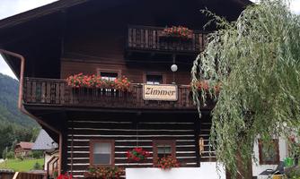A traditional wooden house with a balcony, surrounded by a beautiful green meadow. Colorful flowers adorn the windows and steps.