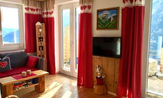 A cozy living room with red curtains, a seating area, and a television. The large windows let in plenty of light and offer a view of the terrace.