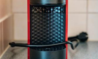 A coffee machine in red and black is standing on a countertop. In the background, power outlets are visible.