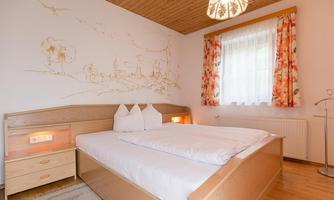 A cozy bedroom with a double bed and beautiful wooden furniture. The room has bright curtains and a mural depicting a rural scene.