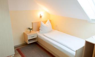 A small, bright room with a single bed and a bedside lamp. The walls are painted in soft colors, and there is a carpet on the floor.