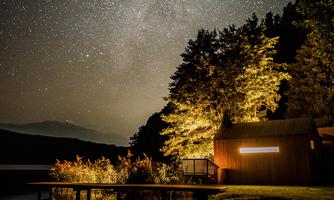 A picturesque landscape at night with a clear starry sky. A cozy wooden house is surrounded by trees and is mirrored in the calm water.