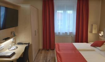 A modern bedroom with a double bed, red curtains, and a television. A desk and a small seating area are also available.