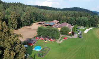 A rural property surrounded by trees with a pool and small red holiday cottages. In the background, meadows and a barn can be seen.
