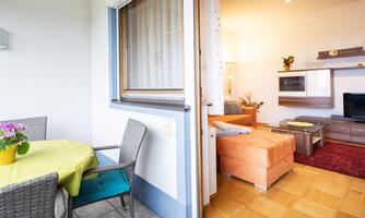 A cozy balcony with a table and chairs, featuring a colorful tablecloth. In the background, there's a bright living room with an orange couch and a television.