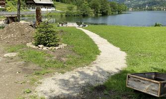 A picturesque path along a tranquil lake, surrounded by trees and mountains. In the background, some cabins can be seen.