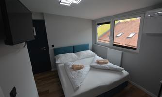 A modern bedroom with a double bed and two towels. Large windows overlooking the buildings outside.
