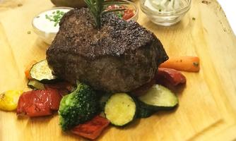 A juicy steak with rosemary on a wooden board, surrounded by colorful vegetables such as zucchini, bell peppers, and broccoli. Accompanied by a baked potato and dipping sauces.