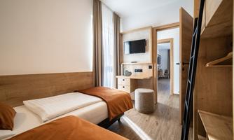 A modern hotel room with two single beds and warm orange blankets. There is a television on the wall and plenty of natural light coming through the windows.