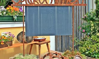A rustic welcome sign stands in the garden, surrounded by colorful flowers and various decorative elements. The background features a friendly house wall.