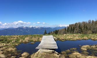 A wooden bridge leads over a waterhole in the mountains. In the background, there are snow-capped peaks and a clear blue sky.
