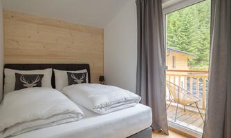 A modern bedroom with a cozy double bed and large windows. The view overlooks a balcony and a green forest landscape.