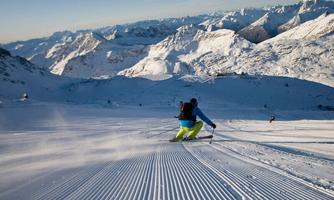 A skier is skiing on a groomed slope in the mountains. The snow-covered peaks shine under a clear blue sky.