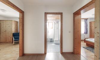 A bright hallway with three open archways. On the left side, there is a room with wooden paneling and a blue chair.