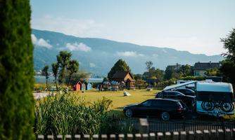 A tranquil landscape by the water with various vehicles and small colorful cabins. In the background, mountains and a clear blue sky can be seen.