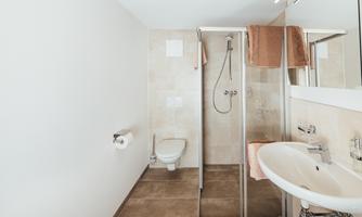 A modern bathroom with a shower, sink, and toilet. The walls are light, and the floor is tiled.
